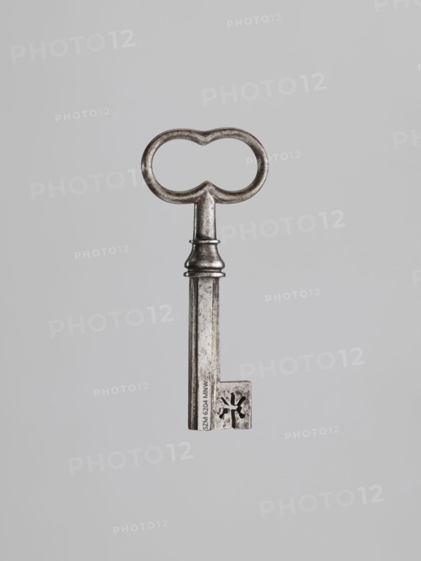 Key, locksmithing. Key with a tubular shaft with a Greek cross cross-section, with a feather slightly thickened toward the edge. Set in a knot with profiles and a vase element. Oval handle, concave at the knot and at the top. 19th century. Height 9.6 cm; width 4.1 cm; depth 1.3 cm., A single metal key is centered against a plain light gray background, showing a rounded bow with two connected loops, a short ornate neck with a ringed collar, and a long rectangular shaft ending in a bit with a cut-out floral-like shape; the metal appears aged with a dull silver-gray color and darker patina in crevices.