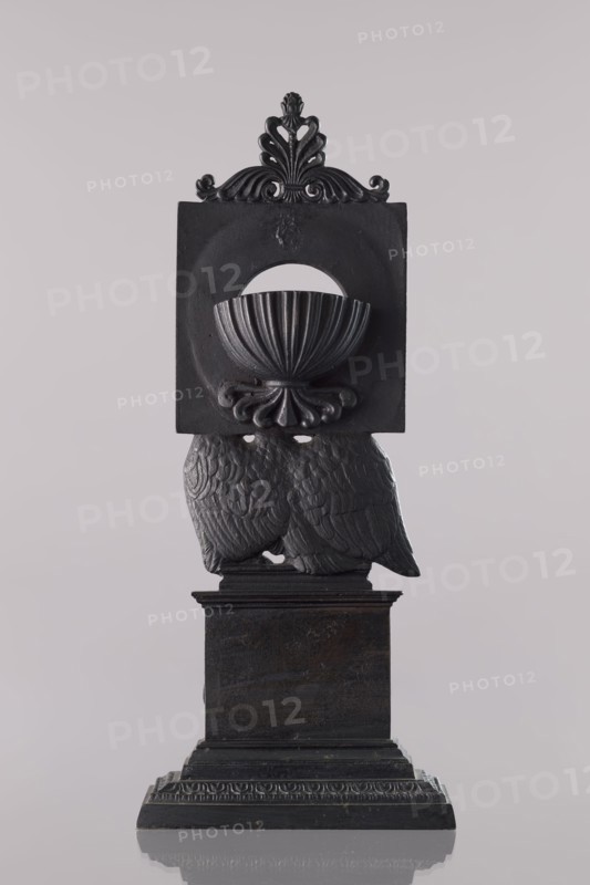 Watch stand with watch, Königlich-Preussische Eisengiesserei in Berlin fl. 1804–1874 ironworks or private foundries; watches; eagle iconography; ca. 1830; Germany; Berlin; varia; height 31 cm, A dark gray sculptural object stands against a pale gray background, featuring ornamental carved details and a square upper section with a semicircular cutout and a shell-like bowl shape, topped by a decorative crest with flowing scrollwork, supported below by a textured pair of wing-like forms with feather patterns, all resting on a stepped rectangular base with carved molding.