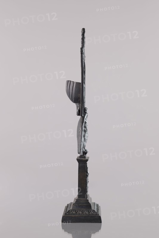 Watch stand with watch, Königlich-Preussische Eisengiesserei in Berlin fl. 1804–1874,  ironworks author or private foundries; watches; eagle iconographic; ca. 1830; Berlin, Germany; varia; height 31 cm, A narrow vertical metal sculpture in dark gray and black tones stands on an ornate square base with stepped tiers, featuring a fluted column rising to a carved human-like figure wearing a tall rounded headdress and a long vertical element extending upward; the sculpture casts a faint reflection on the smooth light gray surface beneath and is set against a pale gray background.
