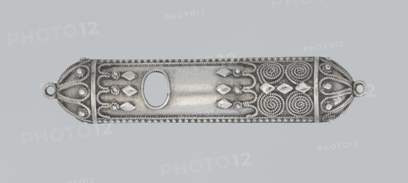 Mezuzah, 19th century, Israel; Judaica; height 0.6 cm, width 6.6 cm, depth 1.3 cm, weight 9 g., A long horizontal metallic object with decorative raised patterns occupies the center of the image; it is primarily silver with darker gray areas accentuating shadows and recessed details. The piece has rounded ends each featuring a small circular loop, and sections of twisted rope-like ridges run along the edges and form spiral motifs near one end. There are several raised diamond-shaped elements and small rounded beads arranged in patterns across the surface, and an oval cutout appears in the central area surrounded by a finely textured border. The background is a uniform pale gray-blue.