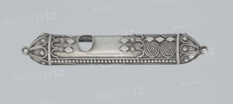 Mezuzah, 19th century, Israel, Judaica; height 0.6 cm, width 6.6 cm, depth 1.3 cm, weight 9 g., A long, narrow metallic object with a silvery-gray appearance rests against a pale gray background; its surface is decorated with ornate raised patterns including twisted rope-like spirals, rounded bead rows, teardrop shapes, and repeating diamond-like raised elements, with smooth polished central areas that show fine surface scratches and an oblong cutout near the middle; both rounded ends feature looped rings and symmetrical rope and bead embellishments, and the overall color palette includes silvery-gray and darker gray tones for shadows and recessed details.