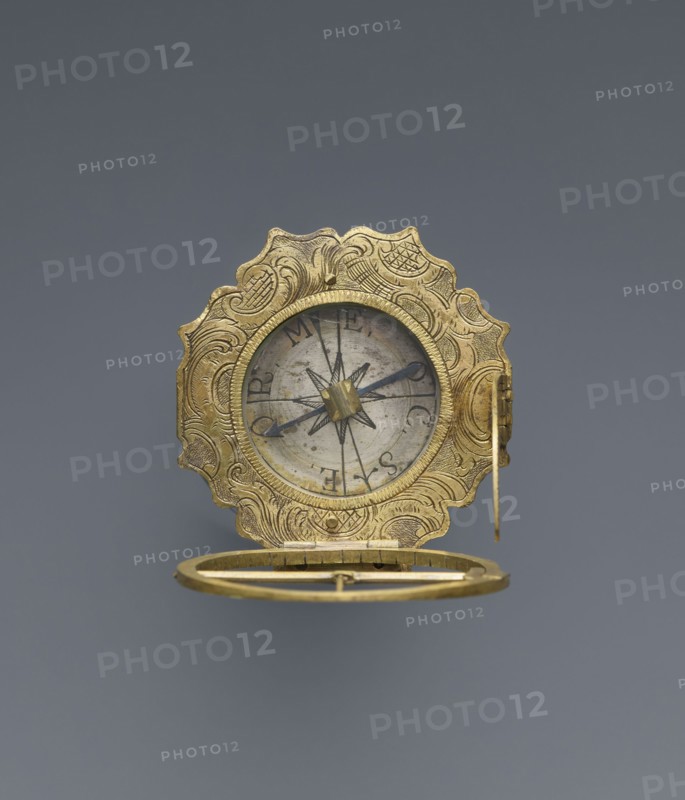 Equatorial sundial by Andreas Vogler ca. 1730–1800, 3rd quarter of the 18th century. Augsburg, Germany. Circular Augsburg-type sundial with a wavy edge; inside the case is a compass with a hinged hour circle and an arched hinged arm marked with geographical latitudes. Engraved decoration on the rim with C-shaped sections and stylized acanthus leaves. Engraved on the underside of the compass case: Eleva. Poli. / Corfu 39 Lyon 45 /' Berlinn 53 Cadiv 36 / Lisabon 39 Paris 48 / Colln 521 Reval 61 / A. Vogler. Function: measuring instrument sundial., An ornate circular metallic object with elaborate scalloped edges and engraved swirling patterns in a golden color, shown open to reveal an inner circular face with a central star-shaped motif and thin dark pointers; the inner face appears silver-gray with aged spots, and the object includes a hinged ring-like component beneath also in gold.