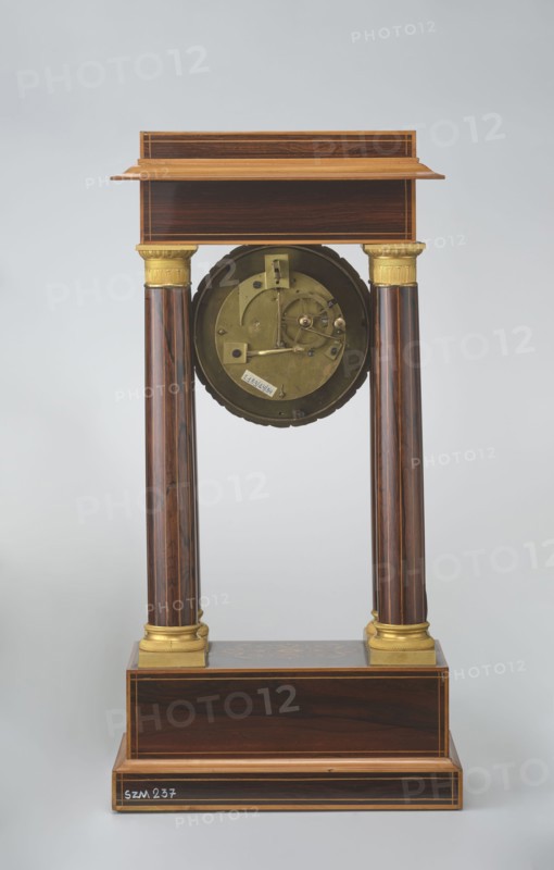 Mantel clock by Alexandre Pertrand fl. ca. 1840. Four-column portico form on a cuboid base and counter-base; boxwood inlays on the front of the cornice and base with a rhombus rosette and a tendril on both pilasters; gilded bronze bases and capitals. Suspended movement with a silver dial with Roman numerals and two keyholes hangs between the columns. Gilded bronze frame with shells surrounds the clock. Oak construction with rosewood facing and light wood insets. Circa 1840, Paris, France. Timepiece; height 47 cm, width 21 cm., A tall wooden structure with a rectangular base and a stepped top supported by a pair of polished vertical columns, each topped with a decorative gold-colored capital and resting on gold-colored bases, with a circular metallic mechanism suspended between the columns; the wood shows dark brown and lighter brown grain with thin lighter inlaid edges, the metallic parts are brass-colored and the capitals and bases are gold-colored, the background is pale gray.
