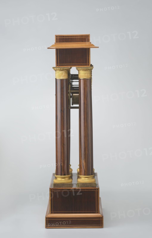 Mantel clock by Alexandre Pertrand fl. ca. 1840. Mantel clock in the form of a four-column portico on a cuboid base and counter-base; boxwood inlays on the front of the cornice and base with a rhombus rosette and a tendril on both stylons; gilded bronze bases and capitals. Suspended movement with a silver dial with Roman numerals and two keyholes hangs between the columns. Gilded bronze frame with shells surrounds the clock. Oak construction with rosewood facing and light wood insets. Ca. 1840, Paris, France. Timepiece; H. 47 cm, W. 21 cm., A vertical wooden structure with a polished dark brown finish and visible wood grain stands against a pale gray background; it has a rectangular stepped base and a small rectangular top shelf in a lighter brown, connected by a group of four tall cylindrical columns with dark brown wood and lighter brown streaks, each topped and bottomed with decorative gold-colored capitals and bases, and some exposed metallic mechanical components visible between t...