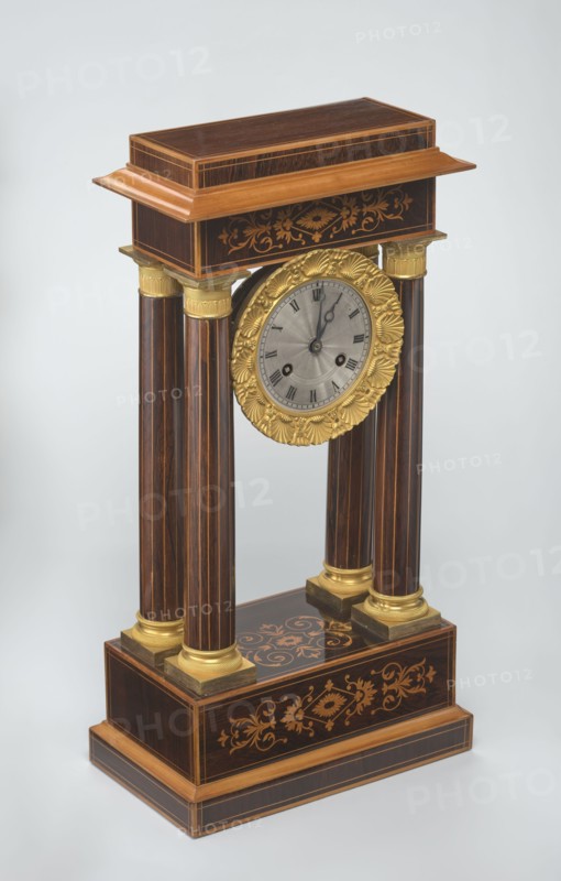 Mantel clock by Alexandre Pertrand fl. ca. 1840. Four-column portico form on a cuboid base and counter-base; boxwood inlays on the front of the cornice and base with a rhombus rosette and tendril on both stanchions; gilded bronze bases and capitals. Suspended movement with a silver dial with Roman numerals and two keyholes hanging between the columns. Gilded bronze frame with shells surrounds the clock. Oak construction with rosewood facing and light wood insets. Ca. 1840, Paris, France. Timepiece; height 47 cm, width 21 cm., A decorative wooden mantel clock with a rectangular base and top connected by four fluted columns, featuring a circular clock face framed by an ornate round gold-colored floral bezel; the wood shows dark brown and lighter brown tones with inlaid floral marquetry in a lighter wood color on the base and top, and gold-colored caps and square plinths at the column ends; the clock face is silver-gray with dark hands, set against a neutral pale gray background.