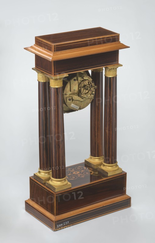Mantel clock by Alexandre Pertrand fl. ca. 1840. Mantel clock in the form of a four-column portico on a cuboid base and counter-base; boxwood inlays on the front of the cornice and base with a rhombus rosette and a tendril on both stiles; gilded bronze bases and capitals. Suspended movement with a silver dial with Roman numerals and two keyholes hangs between the columns. Gilded bronze frame with shells surrounds the clock. Oak construction with rosewood facing and light wood insets. Ca. 1840, Paris, France. Timepiece. Height 47 cm, width 21 cm., A freestanding wooden structure with a rectangular top and base in dark brown wood with lighter brown trim, supported by a group of four tall fluted columns in dark brown with thin lighter streaks and gold-colored capitals and bases, an exposed circular brass mechanism suspended between the columns, and a dark brown plinth with a decorative floral inlay in lighter brown and gold tones, all set against a pale gray background.