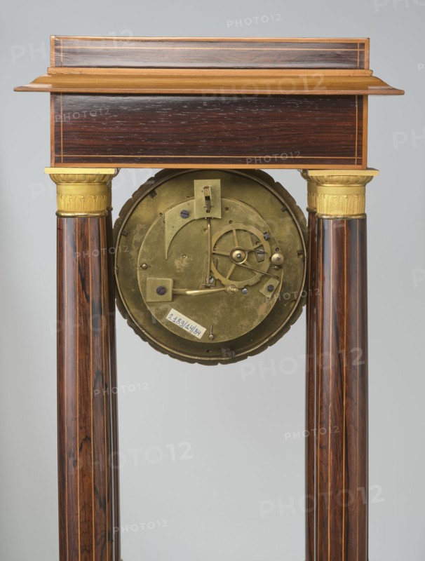 Mantel clock by Alexandre Pertrand fl. ca. 1840. Mantel clock in the form of a four-column portico on a cuboid base and counter-base; boxwood inlays on the front of the cornice and base, with a rhombus rosette and a tendril on both stiles; gilded bronze bases and capitals. A suspended movement with a silver dial with Roman numerals and two keyholes hangs between the columns. A gilded bronze frame with shells surrounds the clock. Oak construction with rosewood facing and light wood insets. Ca. 1840, Paris, France. Height 47 cm; width 21 cm., An upright wooden structure with a rectangular top section in dark brown wood and lighter brown trim, supported by a pair of polished cylindrical wooden columns showing rich brown and reddish wood grain with darker streaks; each column has a decorative gold-colored capital near the top. Between the columns hangs a round metallic mechanism in a muted brass color with visible gears and small fastenings, and a small pale rectangular label attached to i...