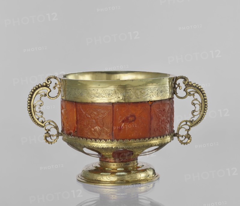Platter with amber plates;, Cylindrical platter on a slightly arched circular foot with oval cutouts at the bottom. Central amber plates decorated with flowers. Upper edge enclosed in a silver hoop engraved with leafy tendril motifs. Sides with scrollwork handles with pearling. 17th century, possibly Królewiec. Garlands and scrollwork ornament. Vessel., An ornate two-handled cup resting on a small round foot, with a metallic rim and base in a yellowish-gold color and a central body made of a translucent reddish-orange material; the handles are intricate and scrolling with beadlike detailing in the same yellowish-gold color, the gold rim and base show engraved decorative patterns, and the reddish-orange central band is carved with relief scenes; the background is a uniform light gray.