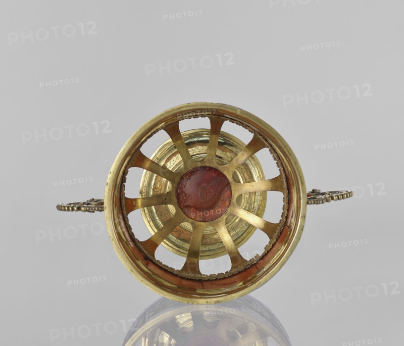 Platter with amber plates, cylindrical platter on a slightly arched circular foot with oval cutouts at the bottom; central amber plates decorated with flowers; upper edge enclosed in a silver hoop engraved with leafy tendril motifs; scrollwork handles with pearling on the sides; 17th century, possibly Królewiec; vessel., A circular metallic object viewed from the front with a ringed outer rim and radial spokes converging on a central disk, showing metallic gold tones and areas of reddish-brown patina, a few ornate handles extending outward on either side with darker metallic details, and a pale gray background with a reflective surface beneath that creates a faint mirrored image.