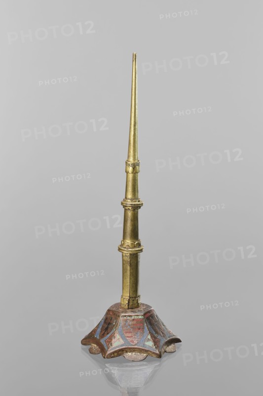 Candlestick, enamel on a hexagonal base tapering toward the top with a shaft; on each side a coat of arms on a blue enamel background eagle and blue lion on horizontal red stripes; between them, below the shields on the edges, rhombuses with engraved dragons. 19th-century shaft; 14th-century Limoges possibly; Barcelona possibly; lighting; height 32 cm, diameter 11 cm., A tall, slender metallic object with a tapered golden shaft rising from a decorative base; the shaft shows a few segmented rings and slight ornamentation and is a metallic gold color. The base is flared and scalloped with a worn painted surface showing muted reds, blues, and hints of green alongside the gold, and it sits on a reflective gray surface that mirrors the base faintly; the background is a uniform pale gray.