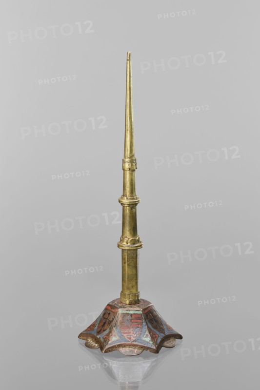 Candlestick, enamel on a hexagonal base tapering toward the top with a shaft; on each side a coat of arms on a blue enamel background eagle and blue lion on horizontal red stripes, between them below the shields on the edges rhombuses with engraved dragons. 14th century Limoges, and 19th-century shaft; lighting. Height 32 cm, diameter 11 cm., A tall, slender metallic object with a tapered upper section in a golden color rises from a decorative base; the base is flared and scalloped with a painted surface showing areas of red, blue, and brown, and the metal shows a slightly aged, patinated appearance; the background is a uniform light gray.