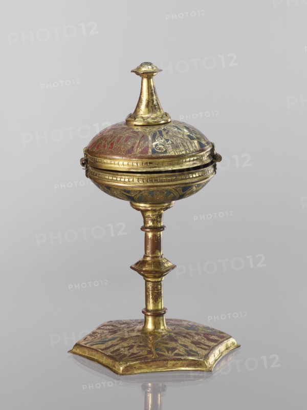 Host ciborium, Copper ciborium on a hexagonal foot, with a smooth shaft with two lacings in the form of a half-cylinder and a hexagonal nodus. The body is a flattened sphere, the upper half serving as a cover; the cover has a conical pinnacle originally topped with a cross. On the foot in the arcades are depictions of the Flagellation, Carrying the Cross, Crucifixion, and Christ in Majesty. On the bowl are busts of angels in circular medallions; on the cover in the trefoil arcades are four depictions of jhakas on the foot. Champlevé enamel in brick red and dark blue, late 14th century, Spain, ritual object; height 28 cm, diameter 16 cm., A gilded metal object with a round, domed top and a finial on its peak, mounted on a slender, segmented stem that rises from a flat, hexagonal base; the surface shows areas of worn gold with decorative painted or inlaid motifs in red, blue, and green, and the piece casts a soft reflection on a neutral gray background.