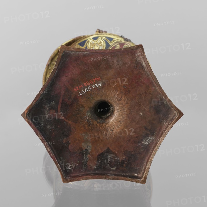 Host ciborium, Copper ciborium on a hexagonal foot, with a smooth shaft featuring two lacings in the form of a half-cylinder and a hexagonal nodus. The body is a flattened sphere, the upper half serving as a cover; the cover has a conical pinnacle originally topped with a cross. On the foot in the arcades are depictions of the Flagellation, Carrying of the Cross, Crucifixion, and Christ in Majesty. On the bowl are busts of angels in circular medallions; on the cover in the trefoil arcades are four depictions of jhakas on the foot. Champlevé enamel, brick red and dark blue, late 14th century, Spain, ritual object, height 28 cm, diameter 16 cm., A close-up view of the bottom of a hexagonal metal object with a central circular hole, showing a worn, textured surface in shades of brown, reddish-brown, dark brown, and hints of black, with areas of lighter tan and small patches of greenish corrosion; the object has a slightly raised rim and beveled edges, and a glimpse of an inner rim decorat...
