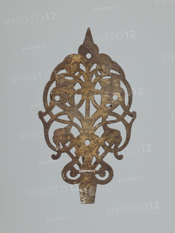 hinge end for a chest, fittings, hinge end from a pair, in the form of an openwork rounded plaque with a tendril motif, two male masks among the twists. Hinge cut off; perhaps originally it was a disc hinge with a sleeve at the end of the plate or a strap hinge, in the form of a clock hand, 1st half of the 17th century, Poland, Central Europe, door fittings, height 21.5 cm, width 12.5 cm, depth 0.2 cm, An ornate, flat metal object with an openwork, symmetrical design is shown against a pale gray background; the metal surface is mottled with brown and dark brown areas and patches of yellowish-gold, and the piece features curved, scroll-like shapes and rounded cutouts with a pointed top and a tapered tab at the bottom.