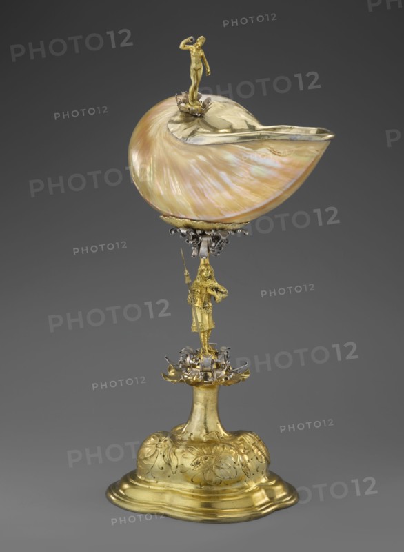 Nautilus Cup. Shell cup on a four-part, profiled foot that transitions into a knobbed foot decorated with flowers. The shaft is a figure of a man holding a halberd, standing on a decoration of volute silver ribbon; the motif is repeated at the base of the cup. Rim framed in silver and topped with a figure of a nude woman with her right hand raised, probably holding a missing element. Case included. Baroque style, masterpiece. 2nd half of the 17th century, Central Europe. Vessel. Height 37 cm; width 17 cm; depth 18 cm. Men; women; shell shape., A decorative object consisting of a large iridescent shell with pearly hues of pink, cream, and green mounted on an ornate metal stem; the shell is partially lined with a shiny golden rim and topped by a small golden human-like figure standing on a floral mount. The stem features a golden human-like figure holding a long staff and stands above a complex floral and scrollwork cluster in mixed metallic tones of gold and silver. The base is a rounde...