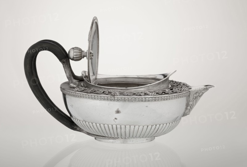 Teapot, Rundell Bridge & Rundell fl. ca. 1788–1845 goldsmith's firm, Scott, Digby & Smith, Benjamin fl. 1802–1807 goldsmiths/sellers; tea vessel, 1803–1804, London, England. Hemispherical teapot on a low cylindrical pedestal; body decorated in lower part with radial curls and at the upper edge with a band of acanthus leaves. Hinged lid surrounded by a raised collar with an applied vine branch with clusters. Double handle of blackened wood set in caps—the upper caps shaped as snake heads, the lower as tails. Applied Czartoryski coat of arms on a paludament with labry and the motto Badz co sie jest. Decoration includes vines, snake iconography, Pogon Ruska coat of arms, rank crowns, princely mitre, mottos, paludamentum, acanthus leaves, clasp, labry. Vessel; height 13 cm, width 24.6 cm, depth 17.2 cm, weight 1002 g., A silver teapot with a dark curved handle sits on a pale background; the teapot has an open hinged lid showing the inside rim and a raised decorative border around the top w...