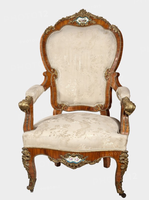 Armchair, origin unknown, neo-Rococo style, with acanthus ornament, flowers, rocaille ornament, fêtes galantes motif, campanulas ornament, and floral bouquets. Neo-Rococo armchair, upholstered backrest, seat, armrests, rose-veneered, decorated with porcelain plaques and cast brass fittings. S-shaped legs widening in the upper section cabriole, arching into a frame with a wavy outline and bulging form; front ends with rollers. Armrests repeat the frame outline, on recessed slightly S-shaped supports topped with volutes. Backrest formed as a symmetrical cartouche, widening upwards, framed by channel sections topped with volutes and with a clearance. Center of front frame and backrest finial have porcelain plaques painted with flowers, framed by a brass frame of rocaille, flowers, and acanthus leaves. Frame edges accentuated with brass strips; brass fittings include a garland at the backrest finial, volutes at the ends of the armrests, acanthus plaques and slippers at the knees and feet. ...