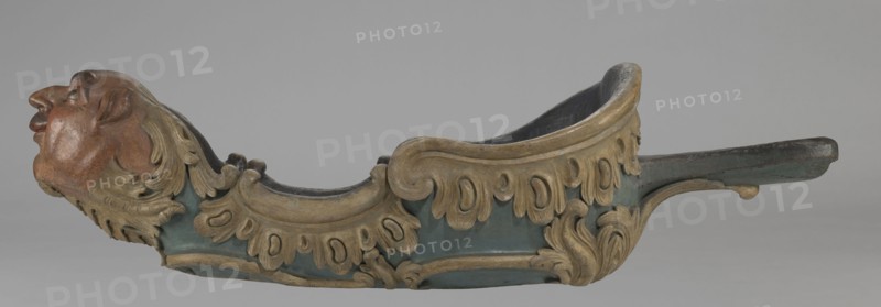 Sleigh, rococo two-seater, carved, polychrome, gilded; 3rd quarter of the 18th century, Poland . Sleigh in the shape of a gondola made using the dugout canoe method, painted blue, with a wavy upper rim highlighted by a half-cylinder and channel sections with rocaille motifs; front in the form of a mascaron painted red, framed with acanthus leaves; rear trestle originally upholstered with carved acanthus underneath; inside the sleds four sockets for connection with skid supports; mascarons, men's heads, rocaille and acanthus ornamentation; height 57 cm, width 208 cm, depth 68 cm., A carved decorative object resembling a boat-like form with a sculpted human head at one end, showing skin tones of reddish-brown and darker brown, and hair carved in a muted gold color; the main body is painted a muted teal or blue-green with ornate carved scrollwork and leaf motifs in shades of gold and beige, and the interior appears darker, near black or deep brown; there are areas of worn dark brown wood ...