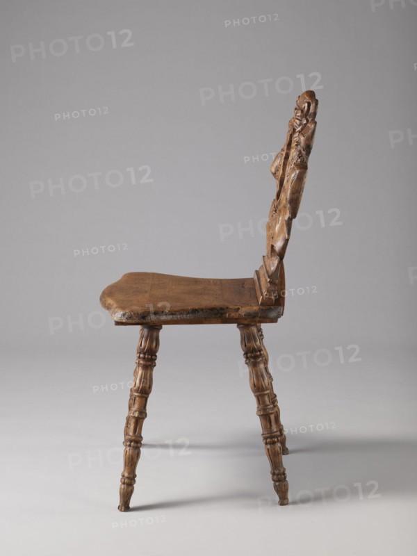 Stool, ca. 1850, possibly Warsaw; neo-Renaissance style; scrollwork and cabochon ornament; piece of furniture — seating piece; height 90 cm, width 44 cm, depth 45 cm., A wooden chair shown from the side with a carved backrest and a flat seat, both in brown wood tones; the backrest features carved decorative shapes and a textured surface, and the seat edge appears worn. The chair has a set of turned legs with carved rings and rounded sections, matching the brown color of the seat and back. The background is a smooth light gray and the floor area is a slightly darker gray, with soft shadows beneath the chair. The visible colors are brown and gray.