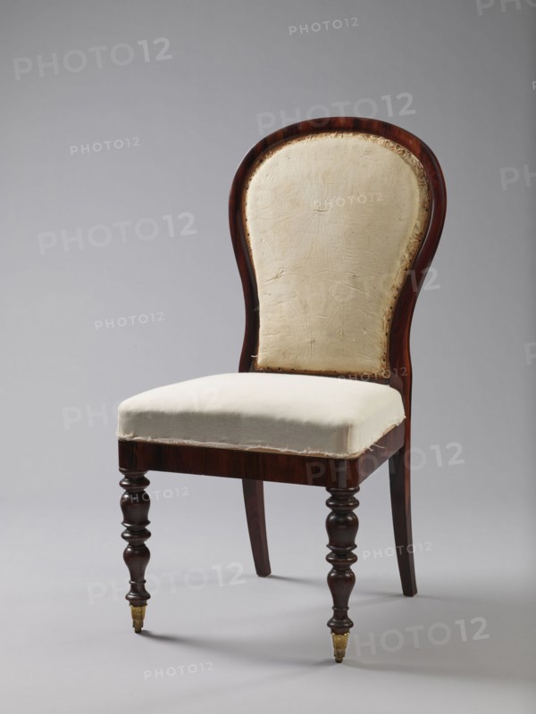 Chair with a padded balloon backrest and a trapezoidal padded seat removable cushion. Front legs are spool-turned, on brass rollers with clamps; rear legs are angular, tapered, slightly arched backwards, continuing into the backrest frames. Low frame with rounded front corners. Upholstery fabric not original: damask with a large floral pattern in purple and cream, finished with upholstery tape. Ca. 1840, Warsaw. Seating furniture; height 97.5 cm, width 48 cm, depth 53.7 cm., A wooden chair with a curved back and a light fabric seat and back panel, the wood showing a dark brown finish with turned front legs and tapered rear legs, the front legs ending in small brass caps, the fabric appearing off-white or cream with visible stitching and worn edges, set against a plain light gray background.