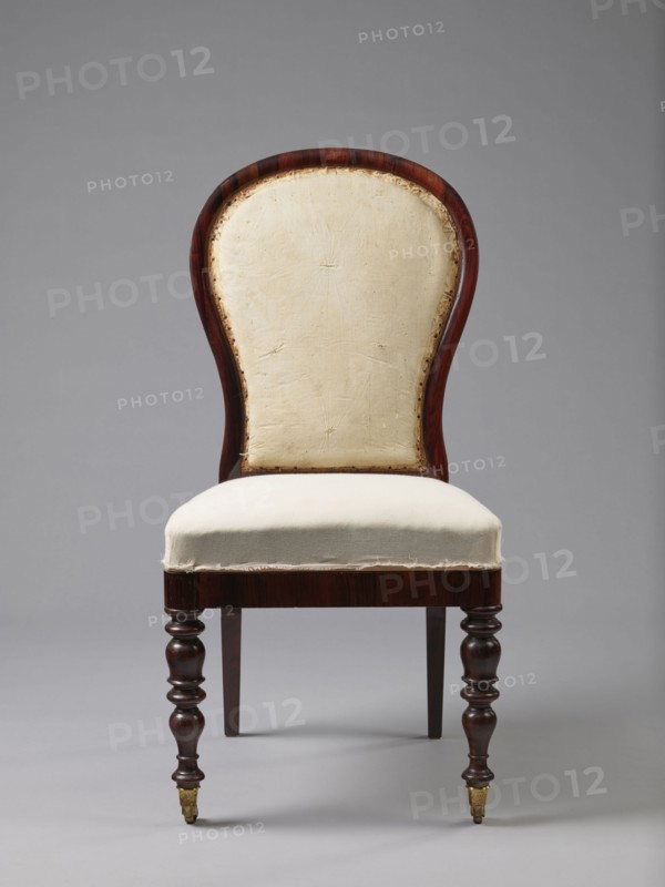 Chair with padded balloon backrest and trapezoidal padded seat removable cushion. Front legs spool-turned, on brass rollers with clamps; rear legs angular, tapered, slightly arched backwards, continuing into backrest frames. Low frame with rounded front corners. Upholstery fabric not original; damask with large floral pattern in purple and cream, finished with upholstery tape. Ca. 1840, Warsaw. Seating furniture; height 97.5 cm, width 48 cm, depth 53.7 cm., A wooden chair with a curved back and upholstered seat is shown against a plain grey background; the wood has a dark brown, reddish tone and a polished finish, the back and seat upholstery are an off-white or cream fabric with visible stitching and slightly frayed edges, the backframe features a shaped outline and exposed inner padding along its edge, the front legs are turned with rounded, carved shapes and end in small brass-colored casters while the rear legs are straight and dark brown.
