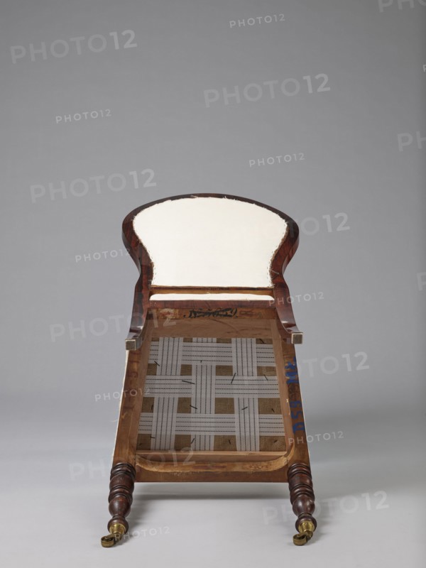 Chair with a padded balloon backrest and a trapezoidal padded seat removable cushion. Front legs are spool-turned, on brass rollers with clamps; rear legs are angular, tapered, slightly arched backwards, continuing into the backrest frames. Low frame with rounded front corners. Upholstery fabric not original, damask with a large floral pattern in purple and cream, finished with upholstery tape. ca. 1840, Warsaw. Seating furniture; height 97.5 cm, width 48 cm, depth 53.7 cm., The image shows the underside of a partially upholstered wooden chair or bench placed against a neutral gray background, with visible wooden frame in dark brown and lighter brown tones, a cream or off-white padded seat area, woven fabric webbing in light beige and off-white forming a lattice pattern, small touches of blue paint on the inner frame, brass or gold-colored caster wheels on turned dark brown wooden legs, and small exposed staples or stitching along the edge of the upholstery.