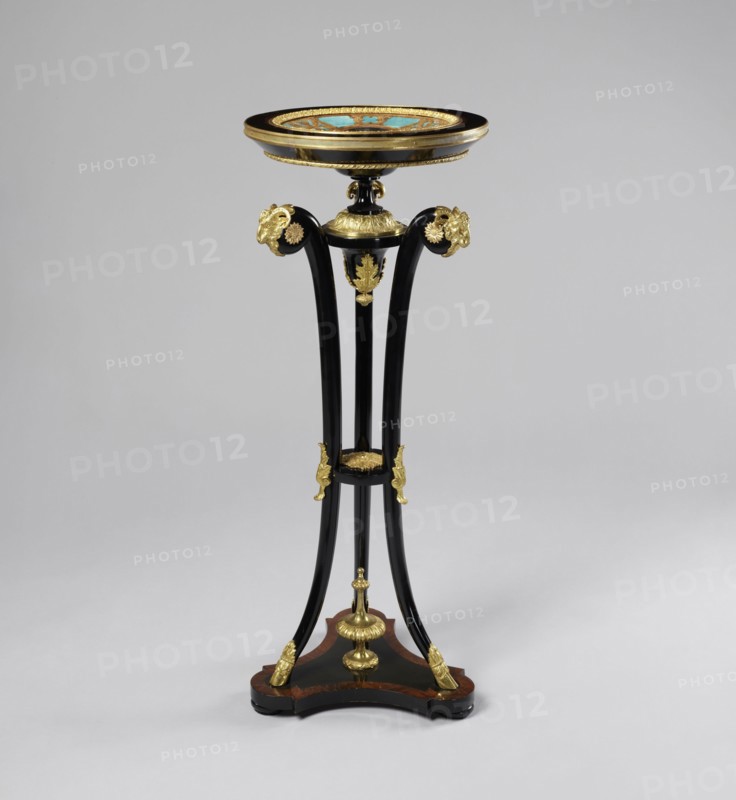 Round table, lacquered black, decorated with gilded cast brass fittings, with a ceramic painted bowl-shaped top supported by three slender channel-shaped supports connected in the middle by a round shelf, mounted on a triangular platform with concave sides. The platform has a profiled rim, with a central pinnacle a spinning top, on three low legs in the shape of strongly flattened spheres. The supports terminate in shoes at the bottom and in sheep's heads at the top. The tabletop is framed in a wooden, profiled frame on a spool-turned support, topped with a pine cone at the bottom and secured by a wooden hoop in the upper part of the supports. The porcelain tabletop plate is painted with overglaze paints, with the gold initial MG under a nine-baton crown in a trefoil field at the bottom, with brown tracery on a blue background border. Decoration includes goat's head motif, plant decoration, crowns, hooves animal legs, and monograms; styles noted: Empire and neo-Gothic. After 1860; like...
