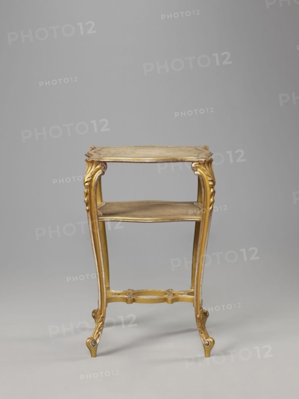 Side table, equipment of representative buildings of the Republic of Poland 1918–1939, plant decoration with acanthus ornament and volutes, neo-rococo style, 3rd quarter of the 19th century, Warsaw; furniture, table; height 77.3 cm, width 50 cm, depth 40 cm, A small ornate wooden table with a gilded finish in shades of gold and muted beige, shown against a plain light gray background; the table has a rectangular top with a slightly scalloped edge, carved decorative apron, and a lower shelf; its four slender cabriole legs are carved with scroll and acanthus motifs and end in small outward-curving feet; a carved stretcher connects the legs near the base and includes a circular decorative element at its center; the overall color palette includes gold, beige, and light brown tones.