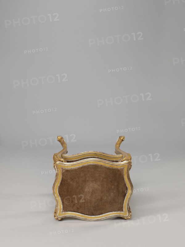 Side table, equipment of representative buildings of the Republic of Poland 1918–1939, plant decoration with acanthus ornament and volutes, neo-rococo style, 3rd quarter of the 19th century, Warsaw; furniture, table; height 77.3 cm, width 50 cm, depth 40 cm, A small ornate stool or low seat is shown from the top edge, with a curved, scalloped wooden frame finished in gold and a muted white trim surrounding a velvet-like upholstered panel in a warm brown tone; the frame has decorative carved handles that curve upward at opposite sides and show gold highlights, and the background and floor are smooth, even shades of light gray.