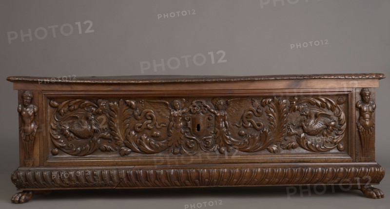 Chest, acanthus twig, fantastic animals, fantastic figures, snakes, coats of arms, herms decorative element, Renaissance style, animal paws, walnut. Chest, cuboid, carved, supported by four low legs, the front ones in the shape of lion's paws, the rear ones angular. On the front, an applied profiled frame enclosing a pseudo-panel carved with grotesque elements on a grained background: in the central section, two figures of winged genii, merging at the bottom into an acanthus twig, holding a laurel wreath above a scrollwork cartouche framing the lock; between the volutes, twigs, snakes and griffins. In the front corners, herms. A prominent plinth molding is symmetrically decorated with gadroons. Straight side walls, originally with doorknobs. Inside, on the left side of the cabinets. Hinges and lock striker made of tinned, engraved steel. Ca. mid-16th century, Italy, piece of furniture, storage unit; height 54.5 cm, width 165.2 cm, depth 57 cm., A carved wooden chest with a rich brown f...