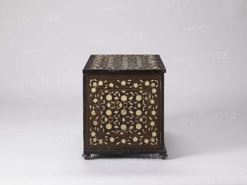 Cabinet, plant decoration, columns architectural, rosettes ornament, early 20th century, India, Europe, piece of furniture, storage piece; height 46.2 cm, width 63 cm, depth 43 cm, A dark wooden box with decorative inlay faces forward, showing an ornate floral pattern in light-colored material against the dark wood on its front and top surfaces; the box sits on small rounded feet and is photographed against a smooth, pale background in shades of white and light gray, with visible colors including dark brown or black of the wood and cream or pale gold of the inlaid floral motifs.