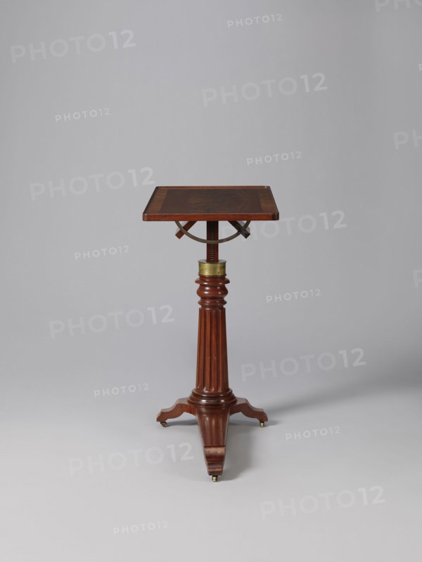 Bedside table, Biedermeier style, mahogany. Composed of an elongated platform outline similar to a triangle on three S-shaped legs with rollers; a fluted shaft mounted on it, tapering upwards, and a rectangular top. The shaft is mounted on the short side of the platform, housing a prismatic column with a metal rack and a top swivel-mounted on it. The height of the top is adjusted using a crank working with the rack. The top can be tilted by a brass ring with a clamp that fixes its position. Turned wooden crank handle. 1815–1830, Europe. Furniture, table. Height 73.2 cm; width 81.2 cm; depth 41.2 cm., A wooden pedestal table with a square top supported by a turned column and a tripod base, showing a reddish-brown wood grain and a small brass band near the top of the column, set against a light gray background with a subtle shadow on the surface.