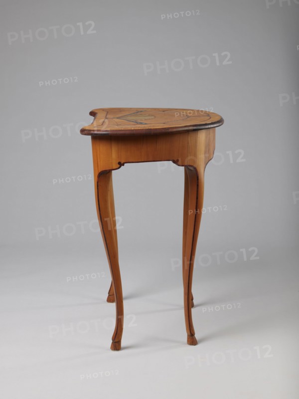 Table, Gift. Pre-war. Kidney-shaped auxiliary table supported by four slender, slightly curved S-shaped legs framed and bound with a rosewood ribbon. Feet in the form of hooves, stained black. Front frame concave with a small central drawer. Tabletop decorated centrally with a fan enclosed by four fields separated by a ribbon of irregular, rounded outline and a narrow crosswise frame. Fan setting inlaid with mother-of-pearl rosettes and bound with tin veining; lower part of the fan features alternating light and dark areas. Tabletop edge veneered with yew solid wood. Mortise lock, single-bolt with brass plate. Circa 1730–1750, possibly Saxony. Height 72.5 cm, width 71.5 cm, depth 46.2 cm., A wooden side table with a warm honey-brown finish stands on a light gray background; the table has a rounded top with darker brown edge trim and a subtle inlaid decorative pattern on the surface, a slightly scalloped apron, and a set of slender, curving cabriole legs that taper to small feet, with v...