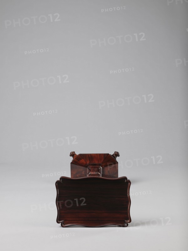 Pedestal table with a rectangular top with a wavy-cut edge and a low wooden gallery, supported by an angular shaft widening toward the bottom, mounted on an octagonal platform on four volute legs. The shaft has lacing in the lower part; the platform is faceted., 1820–1835, England. Furniture; table. Height 75.8 cm, width 39.5 cm, depth 27.5 cm., A polished dark reddish-brown wooden object with a wavy rectangular front face sits on a pale gray surface against a light gray background, showing wood grain patterns and a glossy finish; behind it a matching wooden section with angular facets and small carved rounded protrusions is partially visible in similar dark reddish-brown tones.
