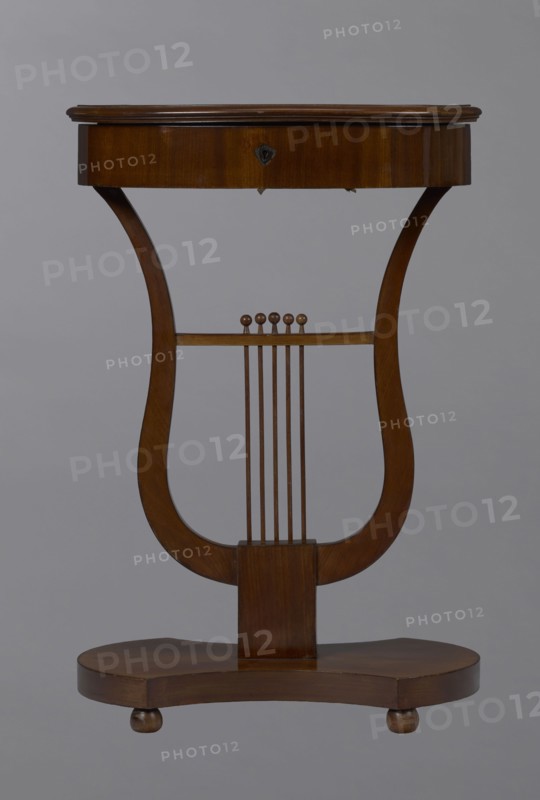 Oval display table with lyre-shaped base mounted on a platform with a concave-convex outline, supported by four spherical legs. Veneered with mahogany. Lyre strings terminate at the top with balls. Edge of the tabletop frame is profiled. Circa 1820–1840, Central Europe. Piece of furniture for displaying objects. Dimensions: height 76.7 cm; width 52.3 cm; depth 37.9 cm., A wooden lyre-shaped table with a rich brown finish set against a neutral gray background; the top is an oval wooden surface with a slightly raised edge and a darker brown hue, supported by curved side supports that form the lyre shape, connecting to a rectangular central post; a horizontal bar across the lyre holds a set of vertical thin wooden rods topped with small rounded finials, all in matching brown tones; the base is a rounded, contoured wooden platform on small rounded feet, also brown.