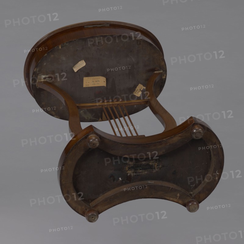 Oval display table with a lyre-shaped base mounted on a platform with a concave-convex outline, supported by four spherical legs. Entirely veneered with mahogany. Lyre strings terminate at the top with balls. The edge of the tabletop frame is profiled. Lyres musical-instrument motif. Piece of furniture for displaying objects. 1820–1840, Central Europe. Height: 76.7 cm; width: 52.3 cm; depth: 37.9 cm. Maker, purchase, A wooden piece of furniture shown from underneath with visible curved legs and a rounded frame in shades of brown, a dark brown underside panel with some lighter brown worn areas, a few small round wooden knobs, several thin wooden slats, and a gray background; visible colors include brown, dark brown, light brown, and gray.