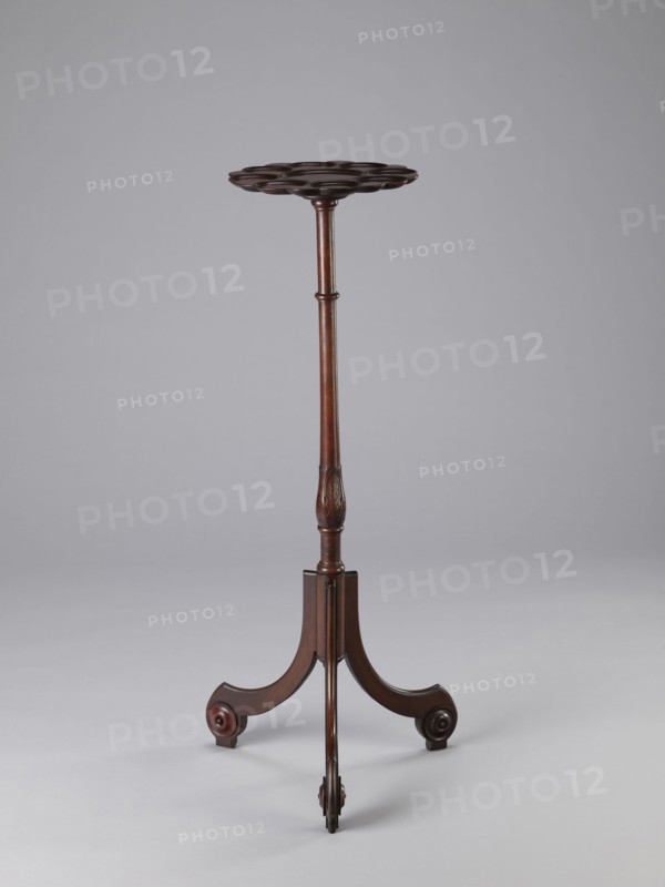 Side table, TABLE with a top in the form of a round saucer mounted on a turned stem forked into a tripod. The top has cut-out recesses for a jug and 8 glasses. The central stem is in the form of a baluster with carved acanthus leaves. Legs made of channel-shaped slats, topped with volutes at the bottom, with turned knobs applied on the sides. 1820–1840, England. Furniture, table. Height 100.5 cm; width 39 cm; depth 39 cm., A wooden stand with a round scalloped top is shown against a plain light gray background; the stand has a slender turned central column with carved details and a dark brown finish, ending in a tripod base with three curved legs, each leg terminating in a rounded foot with circular carved accents; visible colors include dark brown and lighter brown tones on the wood and light gray in the background.