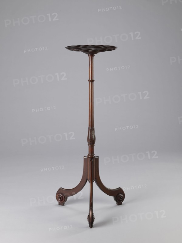 Side table, TABLE with a top in the form of a round saucer mounted on a turned stem forked into a tripod. The top has cut-out recesses for a jug and eight glasses. The central stem is in the form of a baluster with carved acanthus leaves. Legs made of channel-shaped slats, topped with volutes at the bottom, with turned knobs applied on the sides. 1820–1840, England. Furniture; table. Height 100.5 cm, width 39 cm, depth 39 cm., A tall, slender wooden stand with a dark brown finish features a round, scalloped top supported by a turned central column that narrows toward the middle and has carved detailing near the lower section; the base consists of a tripod of curved legs ending in scroll-like feet, all set against a pale gray background.