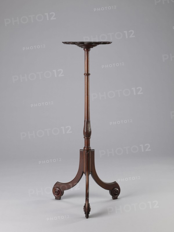 Side table, TABLE with a top in the form of a round saucer mounted on a turned stem forked into a tripod. The top has cut-out recesses for a jug and eight glasses. The central stem is in the form of a baluster with carved acanthus leaves. Legs made of channel-shaped slats, topped with volutes at the bottom, with turned knobs applied on the sides. 1820–1840, England. Furniture; table. Height 100.5 cm; width 39 cm; depth 39 cm., A tall, slender wooden stand with a dark brown finish, featuring a shallow round top with a scalloped edge, a long turned central column with subtle carved detailing, and a tripod base of three curved legs each ending in rounded scroll-like feet; the background is a smooth light gray and the floor area is a slightly lighter gray.