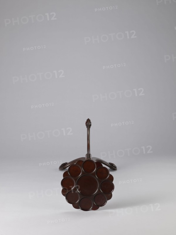 Side table, TABLE with a top in the form of a round saucer mounted on a turned stem forked into a tripod. The top has cut-out recesses for a jug and eight glasses. The central stem is in the form of a baluster with carved acanthus leaves. Legs made of channel-shaped slats, topped with volutes at the bottom, with turned knobs applied on the sides. 1820–1840, England. Furniture; table. Height 100.5 cm, width 39 cm, depth 39 cm., A dark brown wooden object viewed from the end showing a circular flower-like arrangement of round, slightly recessed discs forming a central circle surrounded by a ring of several smaller circles, with a long slender shaft extending back to a curved base that rests on a pale gray surface against a light gray background; visible colors include dark brown, pale gray, and light gray.