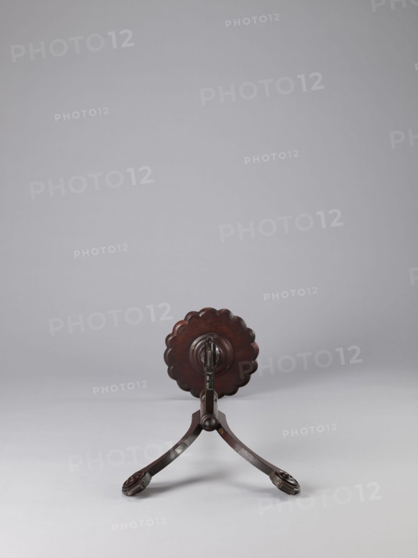 Side table, TABLE with a top in the form of a round saucer mounted on a turned stem forked into a tripod. The top has cut-out recesses for a jug and eight glasses. The central stem is in the form of a baluster with carved acanthus leaves. Legs made of channel-shaped slats, topped with volutes at the bottom, with turned knobs applied on the sides. 1820–1840, England. Furniture; table. Height 100.5 cm, width 39 cm, depth 39 cm., A wooden stand with a dark brown finish is centered on a light gray surface against a pale gray background; the stand has a round scalloped top element and two curved legs ending in carved foot pieces, all in shades of dark brown.