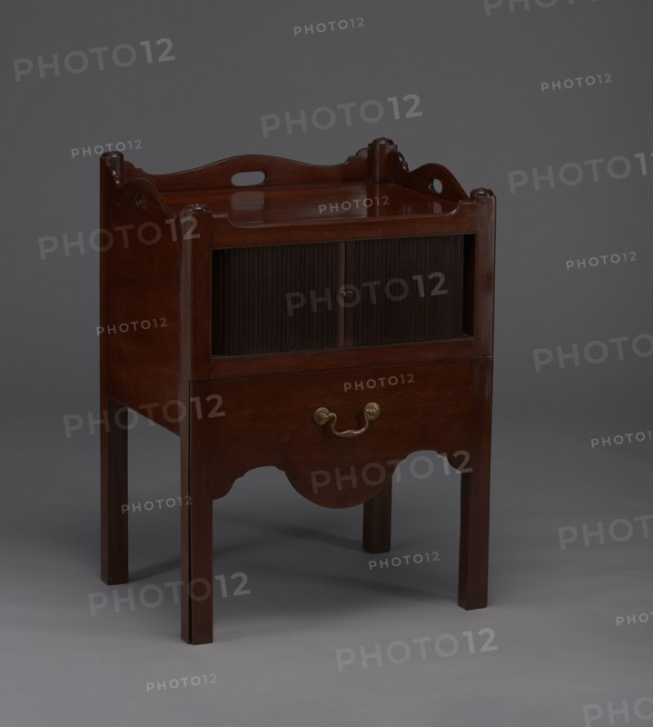 Bedside table with toilet, with a tambour door in the upper section and a pull-out section below with a circular opening for a chamber pot with a lid. Supported by four angular stand-up legs; front legs divided diagonally in half, the front half connected to the front, wavy-cut board of the pull-out section at the bottom. Pull-out handle of gilded brass. Opening cover turned, recessed in the center, with a round handle. Front and side walls of the body have wavy-cut tops with oval openings as handles. A blind consisting of two parts that slide sideways, with small brass knobs. 4th quarter of the 18th century, England, a piece of furniture for hygiene; height 82 cm, width 59.2 cm, depth 45 cm., A wooden piece of furniture with a dark brown finish standing on four straight legs, featuring a rectangular top with curved raised sides and cutout handles, a recessed middle section with sliding tambour-style doors, an open lower drawer front with a brass-colored handle, and a scalloped apron; ...