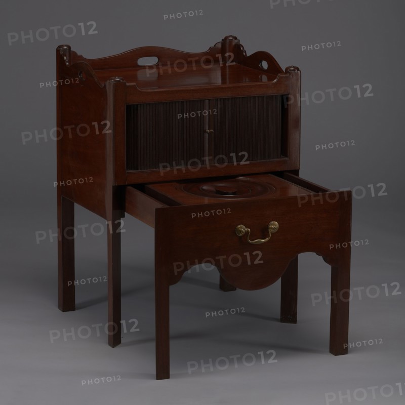 Bedside table with toilet, hygiene, BEDSTAND WITH TOILET, with a tambour door in the upper section and a pull-out section below with a circular opening for a bedside chamber pot with a lid. Supported by four angular stand-up legs; the front legs divided diagonally in half, the front half connected to the front, wavy-cut board of the pull-out section at the bottom. The pull-out handle is gilded brass. The opening cover is turned, recessed in the center, with a round handle. The front and side walls of the body have wavy-cut tops with oval openings as handles. A blind consisting of two parts that slide sideways, with small brass knobs. 4th quarter of the 18th century, England, furniture for hygiene, height 82 cm, width 59.2 cm, depth 45 cm., A wooden piece of furniture with a rich brown finish standing on a group of slender legs, featuring a raised back with curved decorative cutouts and rounded corner posts, a recessed top surface, a pair of vertical slatted panels in the upper section,...