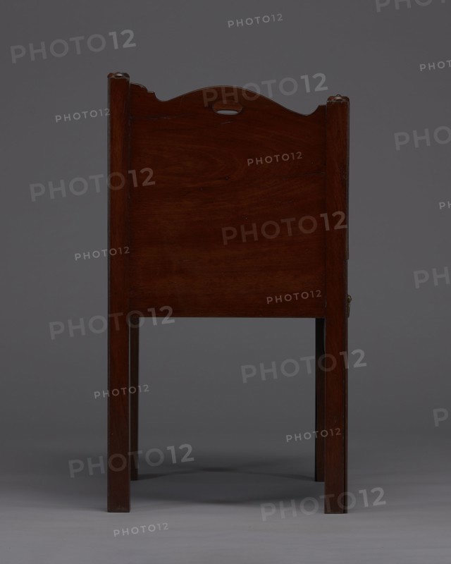Bedside table with toilet, 4th quarter of the 18th century, England. Bedstand with toilet, with a tambour door in the upper section and a pull-out section below with a circular opening for a bedside chamber pot with a lid. Supported by four angular stand-up legs; the front legs divided diagonally in half, the front half connected to the front, wavy-cut board of the pull-out section at the bottom. Pull-out handle of gilded brass. The opening cover is turned, recessed in the center, with a round handle. Front and side walls of the body have wavy-cut tops with oval openings as handles. A blind consisting of two parts that slide sideways, with small brass knobs. A piece of furniture for hygiene. Height 82 cm, width 59.2 cm, depth 45 cm., A wooden chair seen from the back with a dark reddish-brown finish, straight square legs, and a rectangular back panel topped with a gently curved crest that has a small oval cutout; the background is a uniform mid-gray and the floor is a lighter gray, wit...
