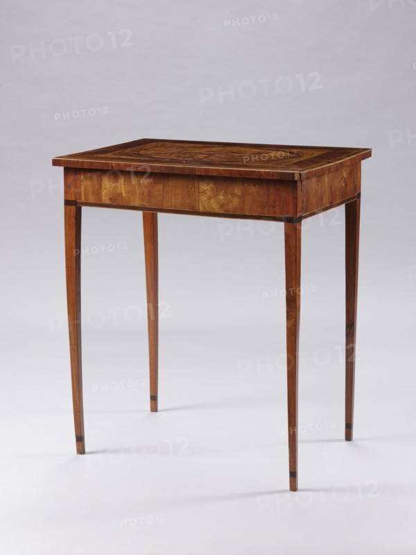 Table, late 18th century, Poland, Classicist rectangular inlaid table supported by four slender, angular, converging legs. On the tabletop, a rectangular field framed by a wide border with light veining, containing an inscribed oval bordered by a wide ribbon and veining. Inside the oval: a seaside landscape with a palm tree on the left, a castle on the right, and two men in Eastern costume standing at a round table with dishes and fruit. Between the S-shaped legs of the table are two bundles. At the bottom of the composition is a cartouche with the initials JJ possibly TT. Figures in Eastern costume, palm trees, seascapes, sailing ships, monograms. Furniture — table. Dimensions: height 73.5 cm, width 63.7 cm, depth 46.3 cm., A wooden table with a rectangular top and slender tapered legs standing on a light surface against a pale background. The table shows a warm brown wood grain with darker brown inlaid details on the tabletop and darker accents near the upper and lower parts of the l...