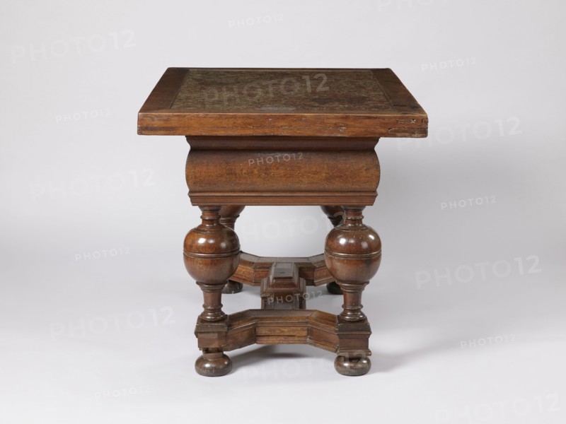 Stock exchange table, provenance unknown, Baroque style, stone tops, rectangular. Rectangular stock exchange table with a stone top enclosed in a wooden frame and a drawer in a high frame, supported by four turned legs joined with an H stretcher made of profiled beams. Baluster legs with massive balls in the upper section, terminating at the bottom with flattened balls. In the central section of the stretcher, a rectangular extension with a pinnacle not preserved. Reinforcing parquetry under the top. Applied drawer bottoms with slides. Stone top polished only on the upper surface. Drawer lock plate in the form of a cartouche topped with a lily. Steel mortise-and-slip lock, double-bolt. Dated to the 4th quarter of the 17th century, possibly Royal Prussia. Furniture, table. Dimensions: height 88.5 cm, width 121.3 cm, depth 90.4 cm., A wooden table with a rectangular top and a carved apron sits against a plain light gray background; the table is brown with darker brown grain and worn area...