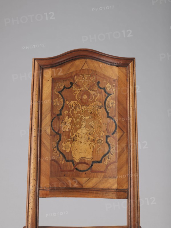 Chair, origin unknown, Rococo style, ca. 1750–1765, Germany. Rococo chair with volute and shell ornamentation, plant decoration including acanthus, female figures, flowers, and ribbon-and-pincer motifs. Boarded inlaid high backrest showing a lady in a crinoline surrounded by flowers, framed by two intertwining U-shaped ribbons. Rectangular originally upholstered seat supported by cabriole legs decorated in bas-relief with motifs at the top shells on the knees terminating in volute feet. Wavy-cut lower frame decorated with acanthus leaves; seat frame with wavy outer outline enclosing a padded central section. Backrest has a clearance, angular stiles, and a wavy-curved upper section. Piece of furniture seating, height 115 cm, width 53 cm, depth 63 cm., A wooden chair back with a curved top and a rectangular inset panel featuring a decorative marquetry scene; the wood shows warm brown and honey tones with darker brown grain and black inlaid accents; the central inlaid design depicts a sea...