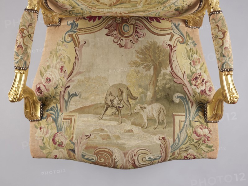 Armchair, attributed to Jean-Baptiste Oudry 1686–1755 and an unknown co-creator; carved, gilded, and upholstered Rococo armchair in the à la reine style. Tapestry upholstery Aubusson and/or Beauvais? featuring scenes from Jean de La Fontaine's fables framed with flowers: backrest depicts an insect nest in a landscape illustration of The Hornets and the Bees, seat depicts The Wolf and the Lamb by a stream. Decorative motifs: wolves, lambs, insects, flowers, bees, plant decoration, shells, rocaille elements, acanthus leaves, bunches of roses. Form: roughly rectangular backrest with slightly concave side rails, convex top and rounded corners; trapezoidal seat with slightly bulging frames; recessed, volute-topped armrests on U-shaped supports; S-shaped cabriole legs with acanthus-shaped volute feet. Function: seating piece. Date: 3rd quarter of the 18th century. Place of origin: France. Dimensions: height 106.5 cm; width 77.5 cm; depth 79.4 cm., A decorative upholstered chair seat seen fro...
