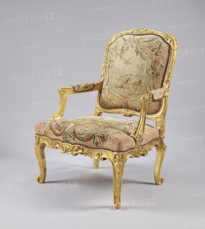 Armchair, maker, upholstery fabric possibly Aubusson or Beauvais; tapestry illustrations after Jean-Baptiste Oudry 1686–1755 and Jean de la Fontaine 1621–1695 fairy tales; motifs include flowers, vultures, wolves, birds, pigeons, wells, shells; Rococo ornament and style; cabriole legs; third quarter of the 18th century, France; seating piece of furniture; height 107.3 cm, width 76.8 cm, depth 77.5 cm., A gilded wooden armchair with ornate carved frame in gold, upholstered with a tapestry fabric showing floral and scenic woven patterns in shades of pink, beige, green, blue, and cream; the seat, back, and arm pads are covered in the patterned fabric edged with a row of small dark studs; the chair has curved legs and scrolled armrests with carved gold details, all set against a neutral light gray background.