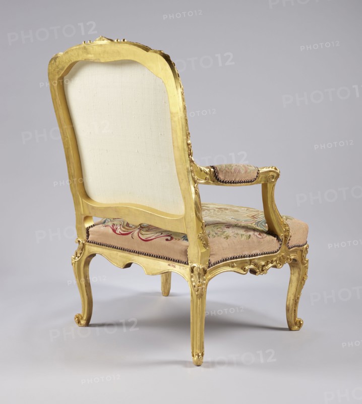 Armchair, upholstery after Jean-Baptiste Oudry 1686–1755 designs; co-creator, tapestry/ upholstery woven in Aubusson and/or Beauvais, France. Upholstery illustrates scenes and motifs from Jean de La Fontaine’s fairy tales 1621–1695: fairy-tale scenes with wolves, vultures, birds and pigeons, animals, wells, flowers, shells Rococo ornament, Rococo style, cabriole legs. Third quarter of the 18th century. Seating piece of furniture; height 107.3 cm, width 76.8 cm, depth 77.5 cm., The image shows the back and side of an ornate upholstered armchair with a carved wooden frame finished in gold. The chair has a tall back with a plain light-colored fabric panel, curved arms with padded armrests covered in patterned fabric, and a patterned seat cushion in muted tones. The wooden frame features decorative carved details on the legs, apron, and arm supports, and the legs end in curled feet. Visible colors include gold on the frame, light beige on the back panel, soft pink and muted green in the pa...